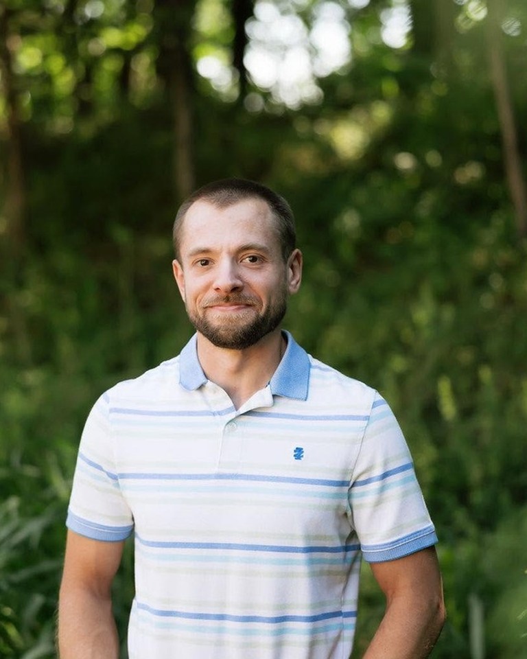 Portrait of Zach Noell outdoors in a striped polo with greenery behind him